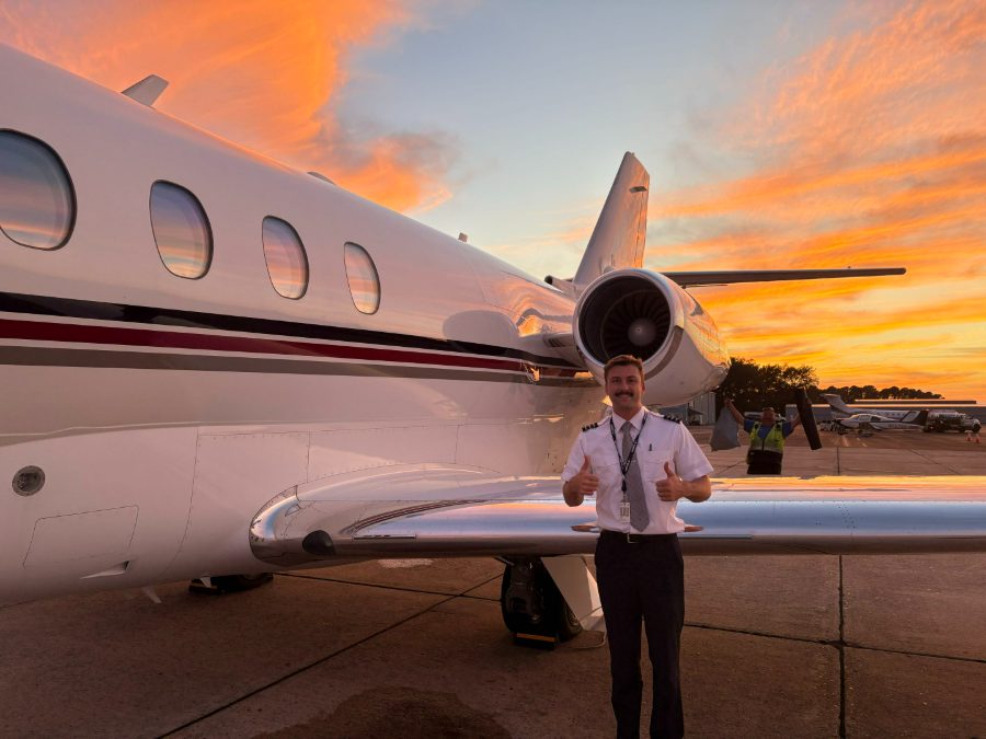 Ethan Pettit in pilot’s uniform, giving a thumbs up beside the business jet he flies, on the apron at sunset.